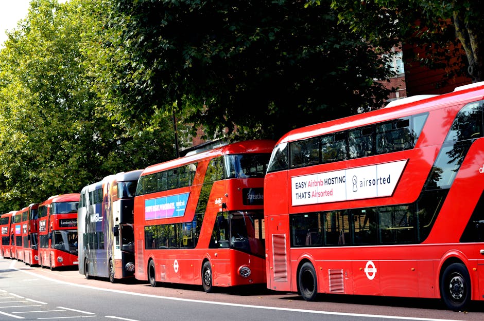 A row of six red double-decker buses parked on a street next to a sidewalk, with various advertisements visible on the side panels of the buses. The buses are aligned parallel to each other, with some