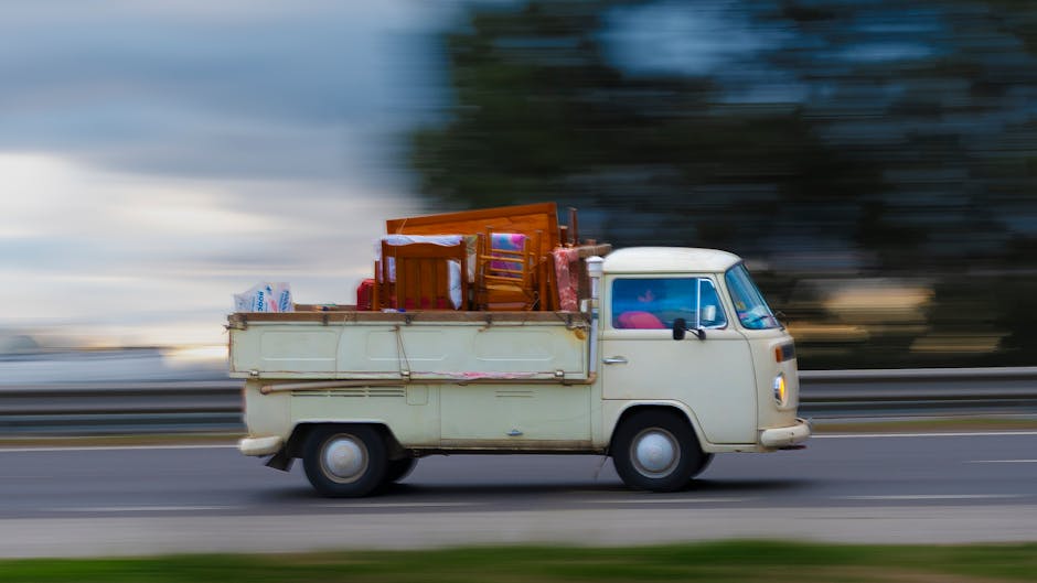 A vintage cream-colored van is shown moving along a roadway during daytime, loaded with various household furniture and packed boxes positioned in the cargo area. The furniture includes wooden chairs